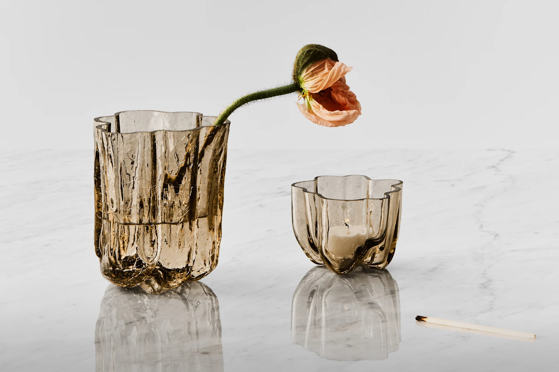 To teksturerede ravfarvede glasbeholdere, den ene med en ferskenfarvet valmue, den anden et tændt lys, på hvidt marmor ved siden af en brændt tændstik.