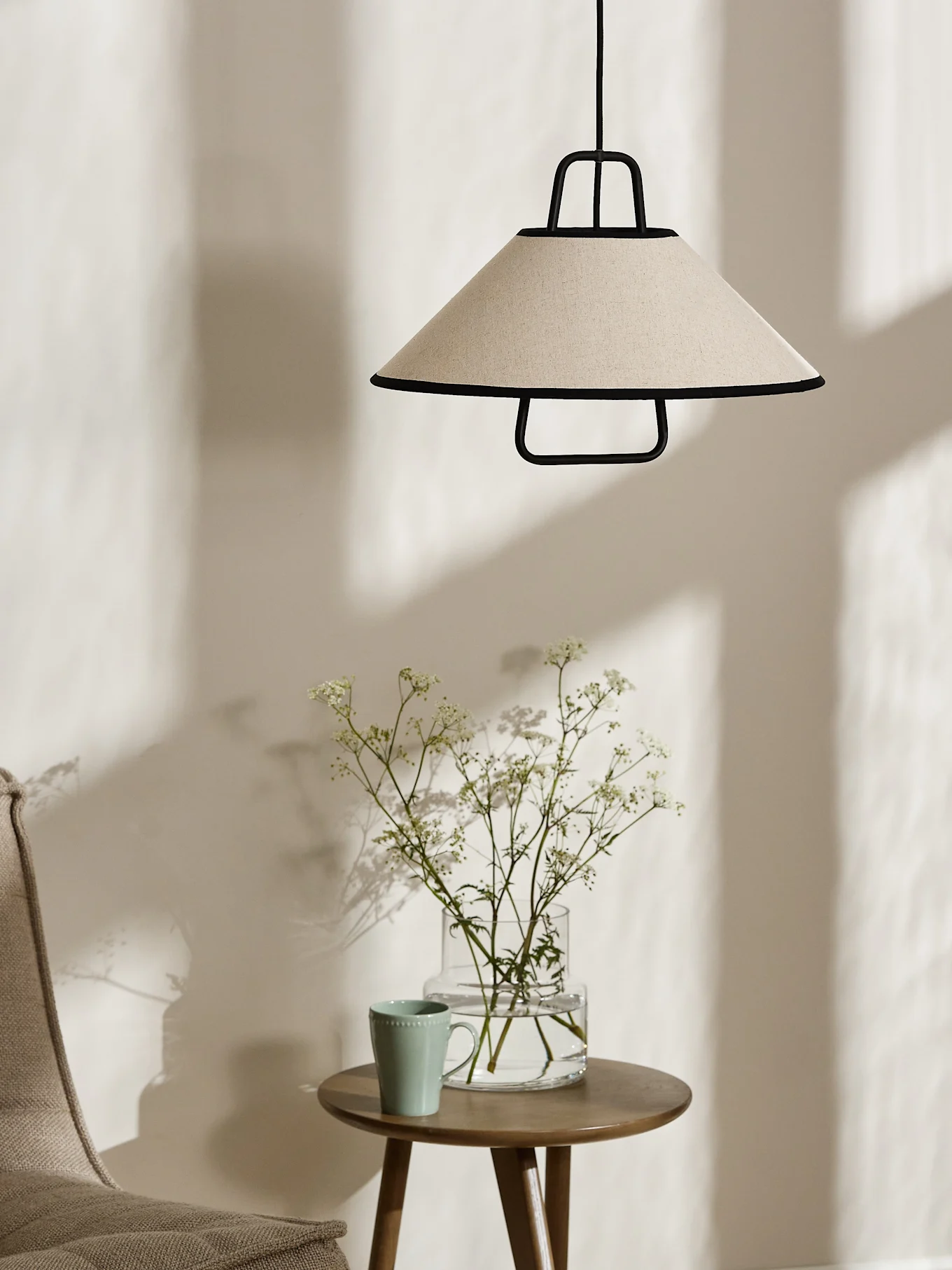 En konisk cremefarvet pendellampe med sort kant hænger over et træsidebord med en vase hvide blomster og et lysegrønt krus.