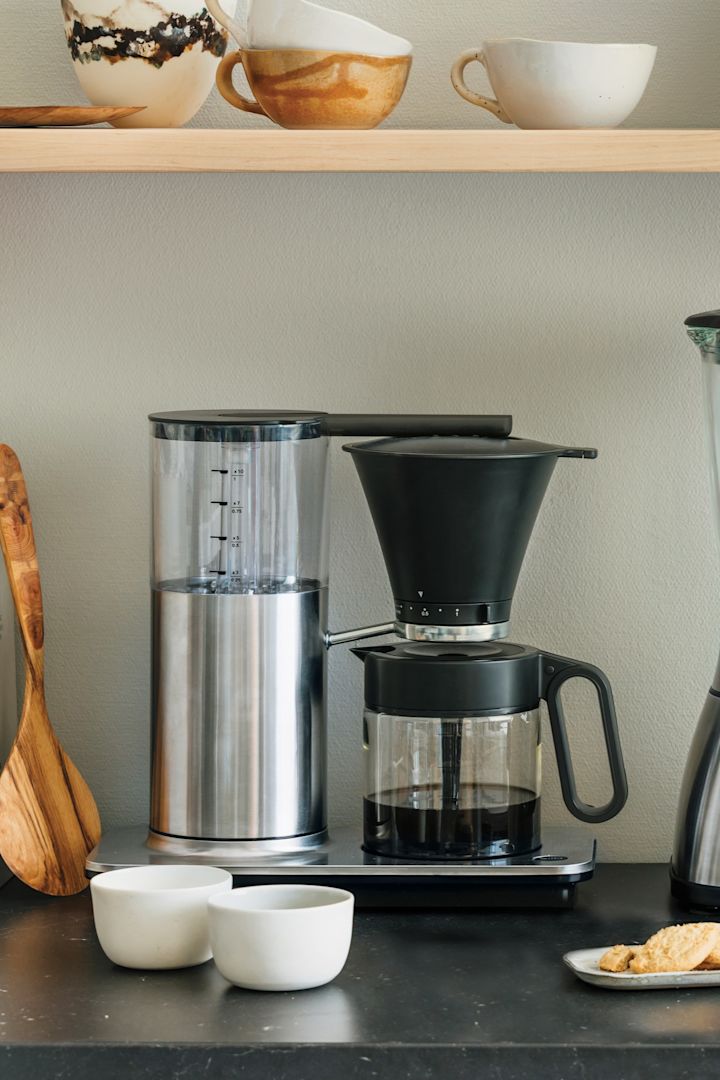 Du har også brug for de rigtige apparater til din hjemmekaffebar. Her kan du se en klassisk Wilfa-filterkaffemaskine på et køkkenbord med en hylde med skåle og en brødrister over.