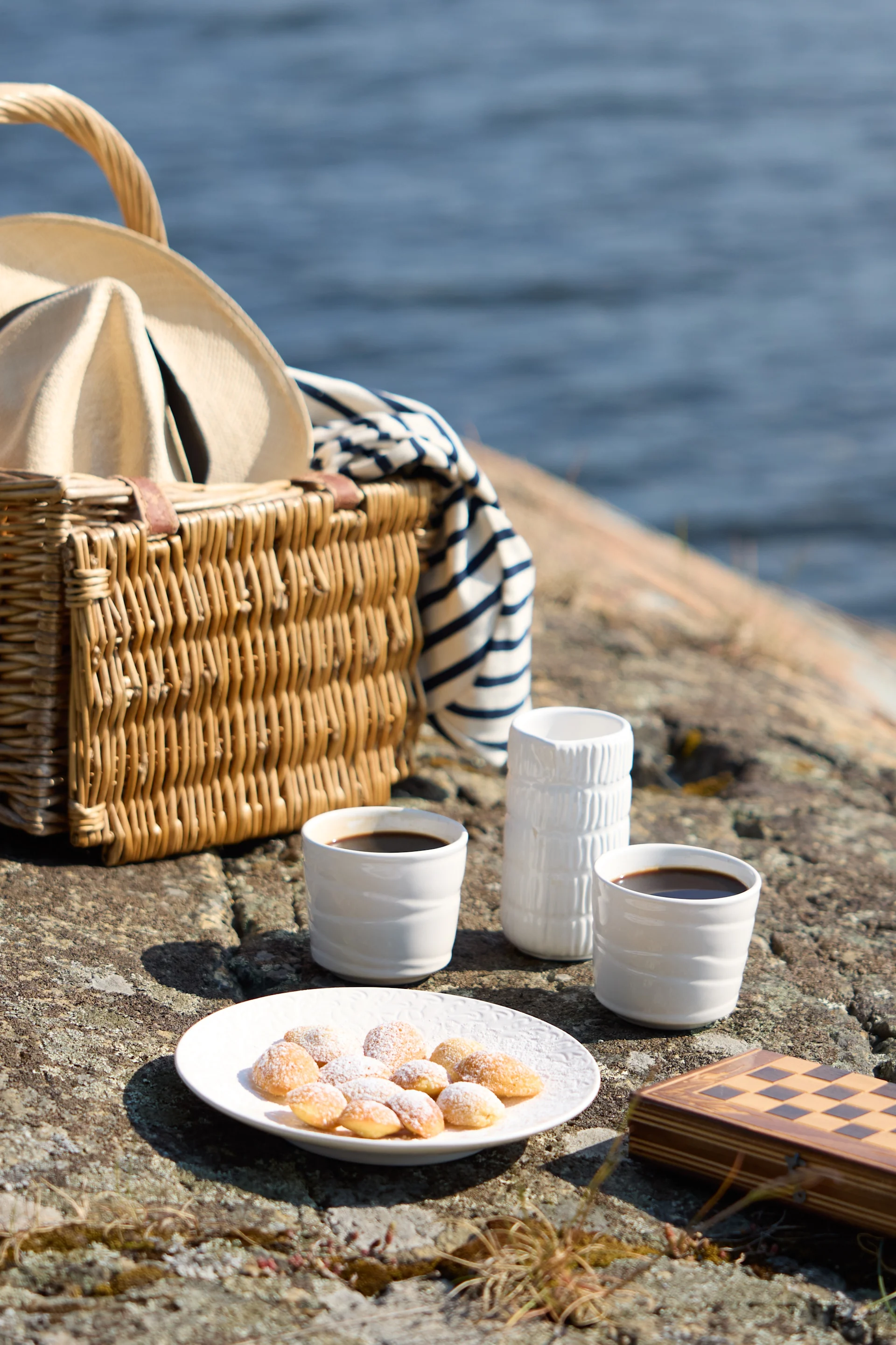 Et picnicarrangement på klippefyldt terræn nær vand, med kaffe, kager og et skaksæt.