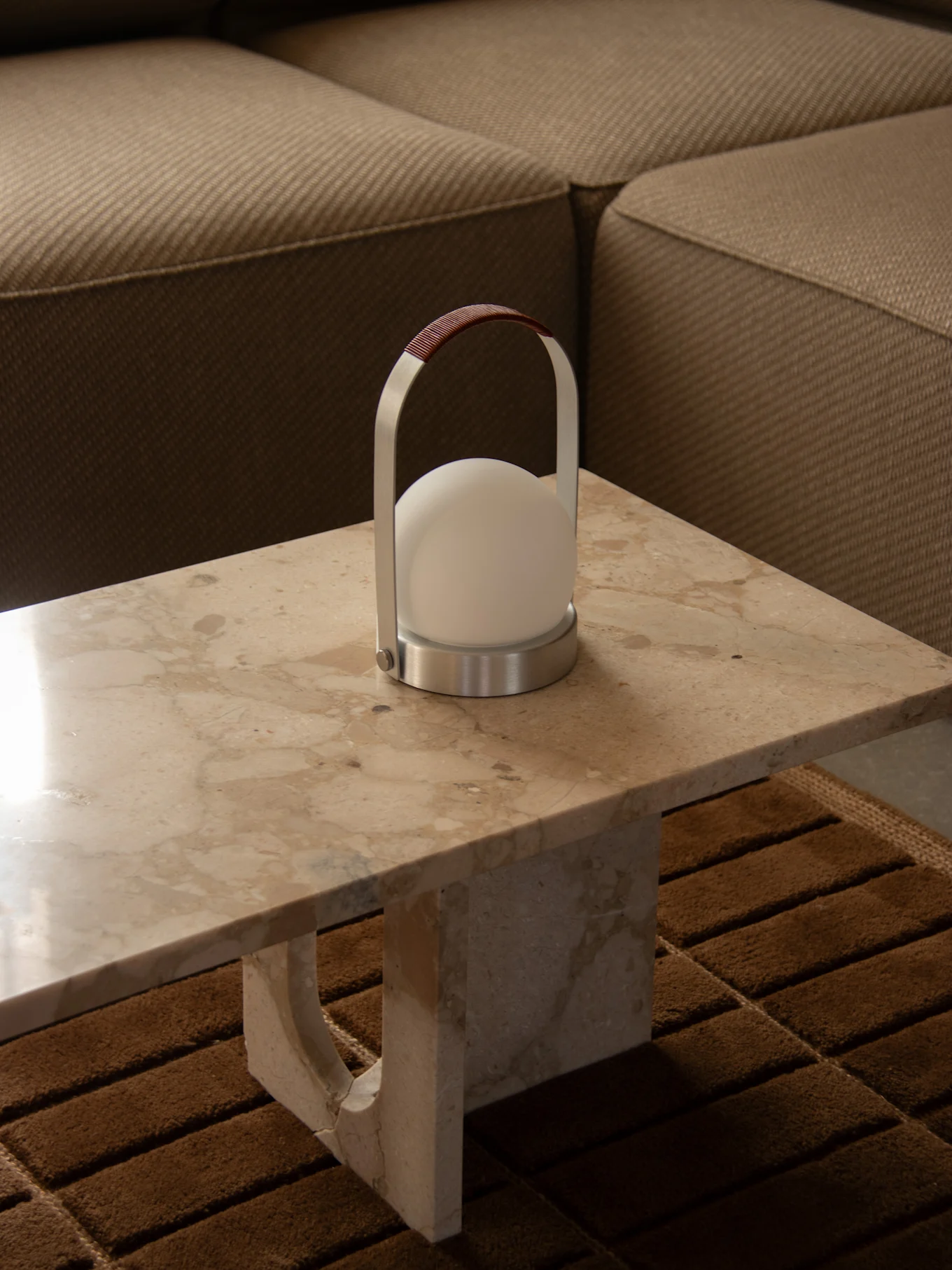 Moderne bærbar lampe med en sfærisk hvid lys, sølvramme og brunt læderhåndtag på et marmorbord ved siden af en sofa.