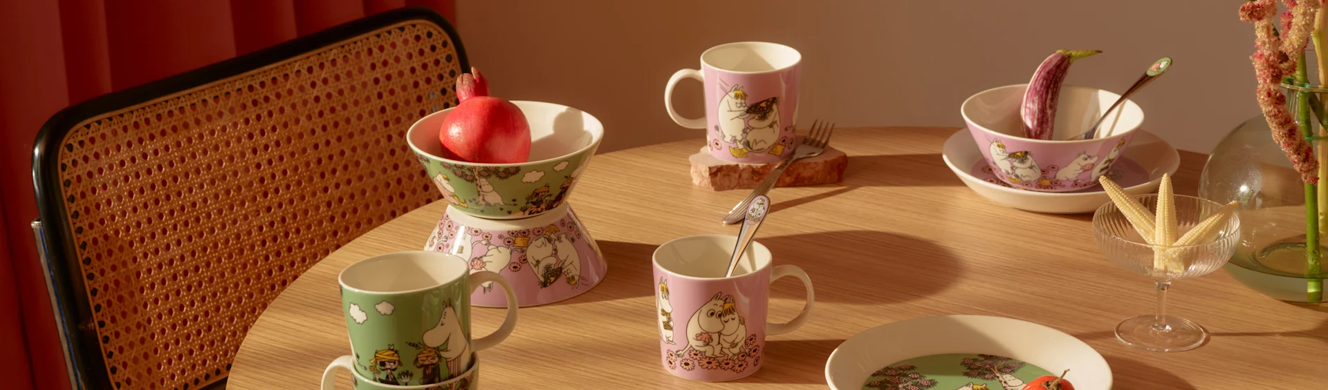 Et træbord dækket med grønne og lyserøde Moomin-krus, skåle og tallerkener, granatæble, aubergine og minimajs.
