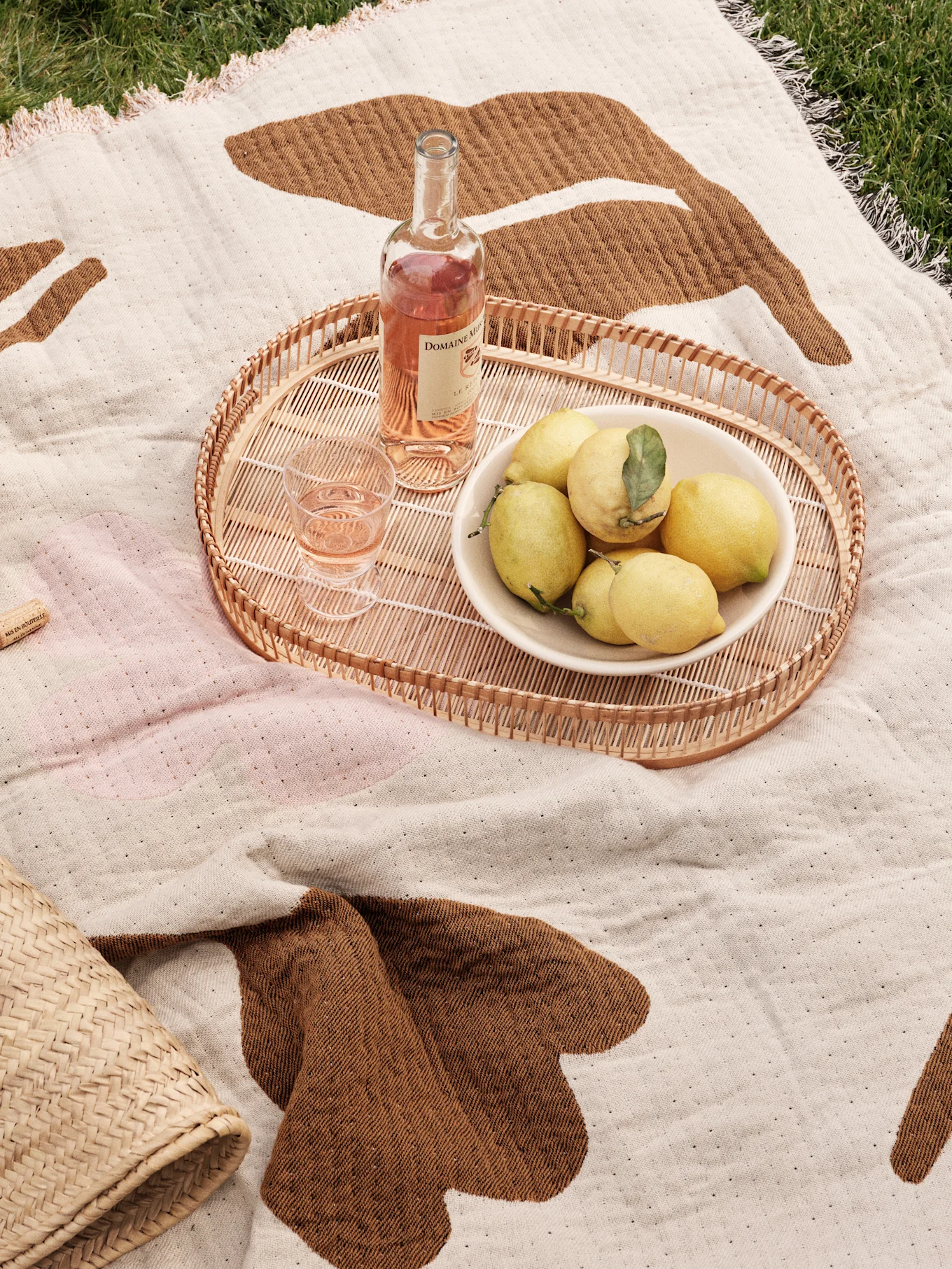 Picnictæppe på græs med flettet bakke, rosévin, glas og en skål citroner.