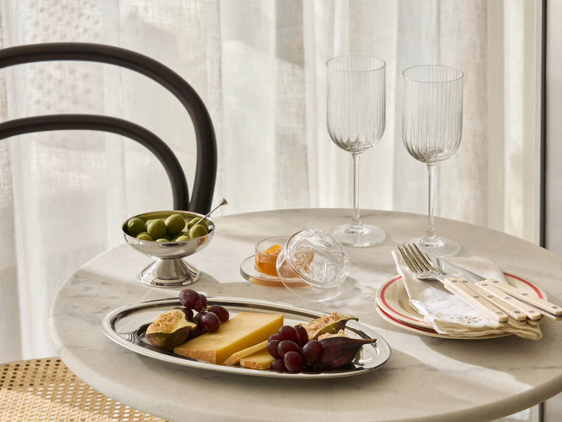 Et marmorbord dækket med et sølvfad med ost, vindruer, figner, en skål oliven, syltetøj, to riflede glas og bestik på tallerkener.