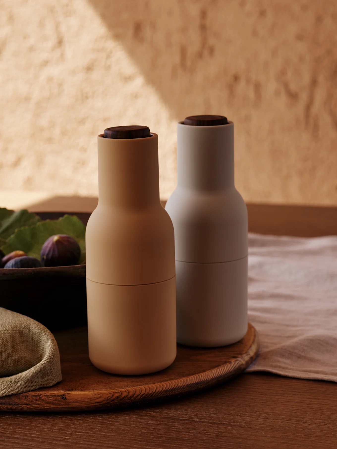 To moderne krydderikværne med trælåg, en beige og en lysegrå, på et rundt træfad med figner.