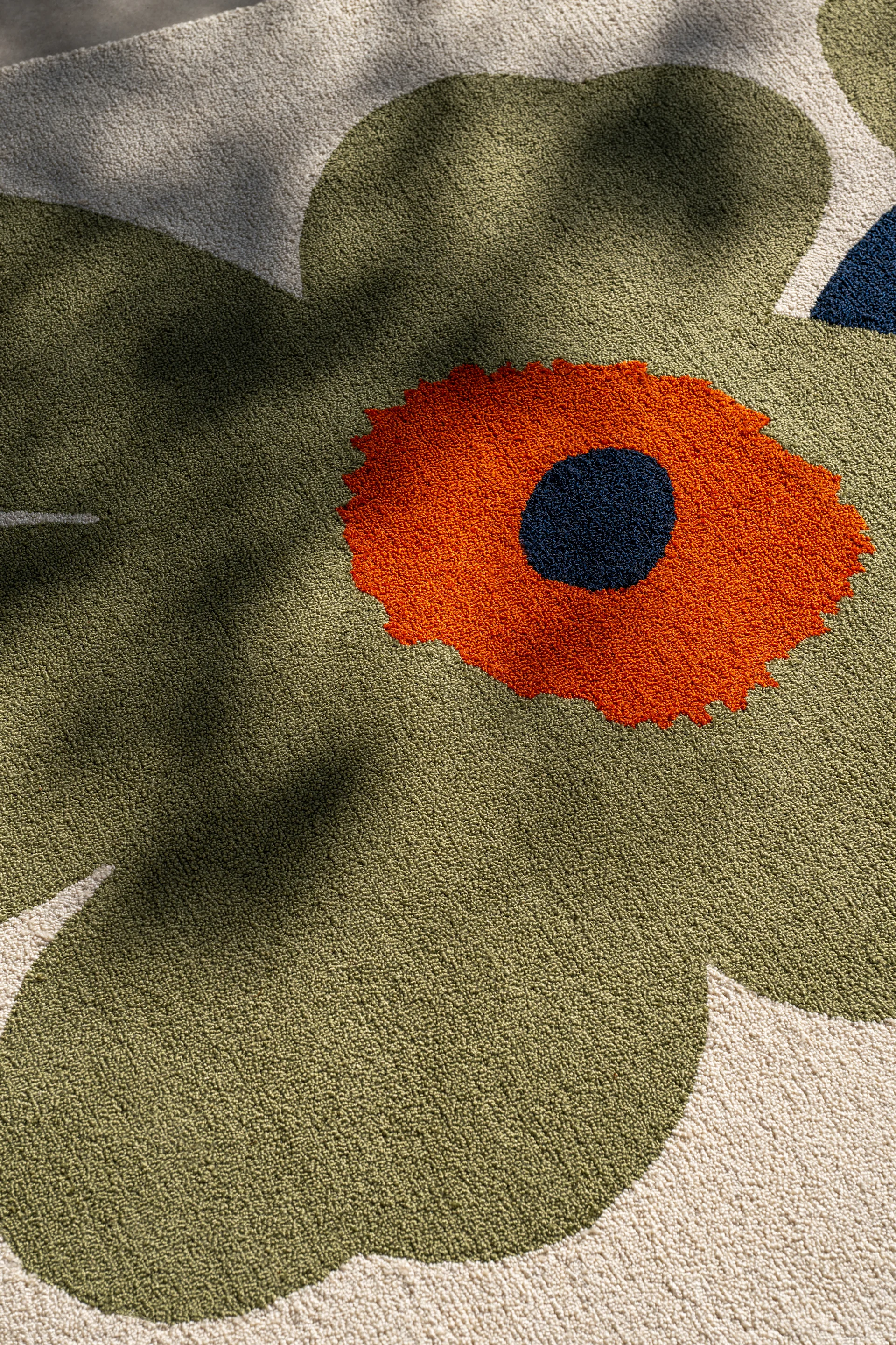 Nærbillede af et tæppe med et stort, stiliseret blomsterdesign. Olivengrønne kronblade, orange midte og en mørkeblå prik på cremefarvet baggrund.