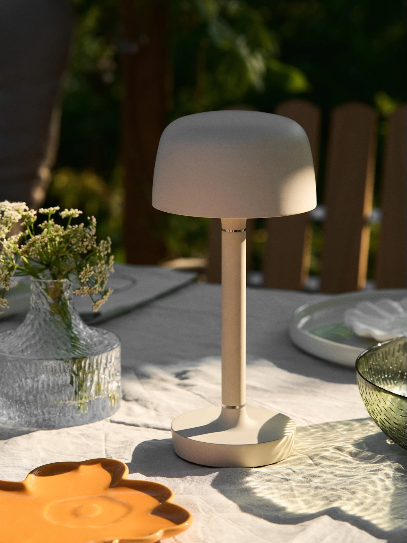 Beige bærbar bordlampe på et udendørsbord med blomstervase og plettet sollys.