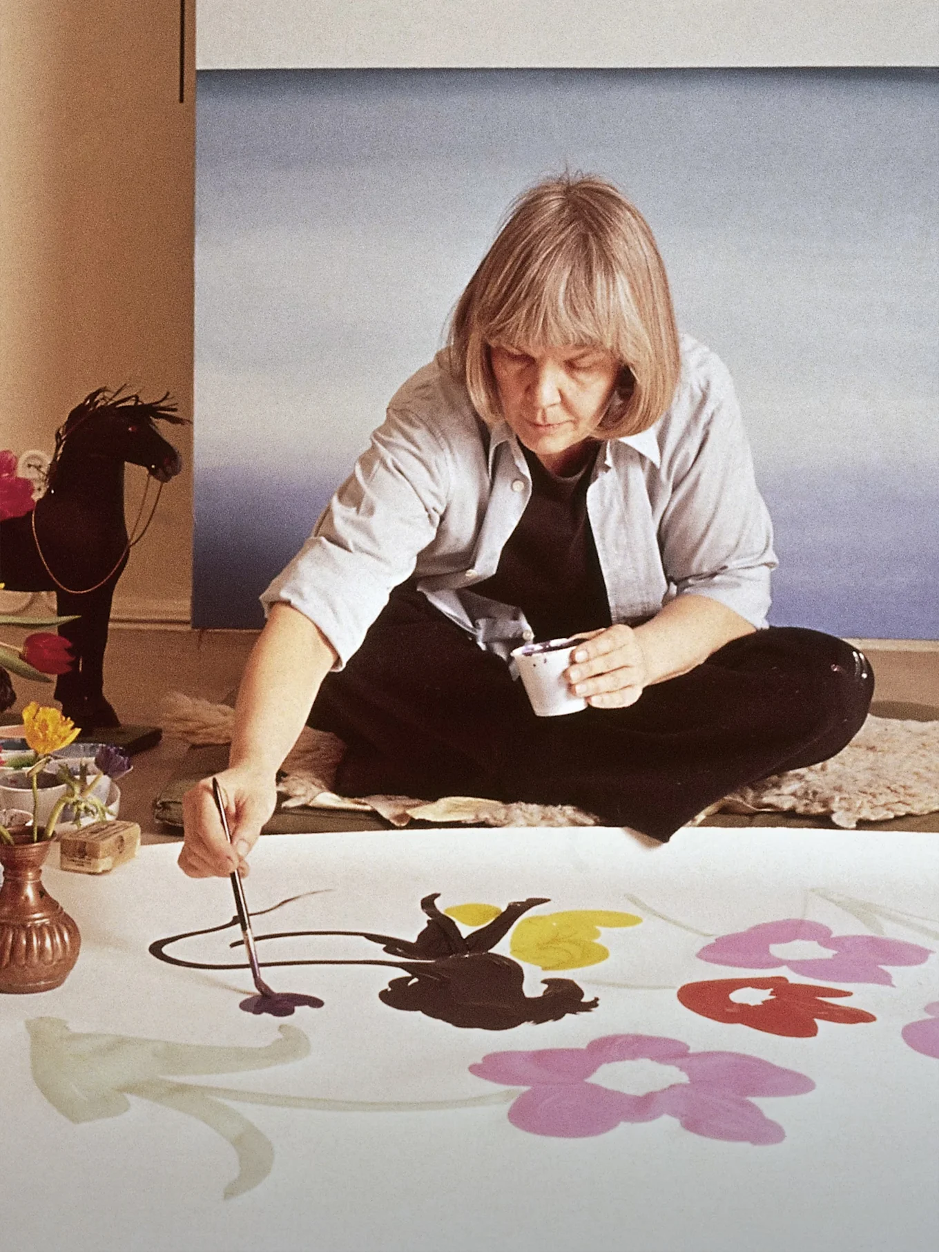Kunstneren Marimekko Maija Isola sidder på gulvet og maler store, farverige blomster på et hvidt stof.