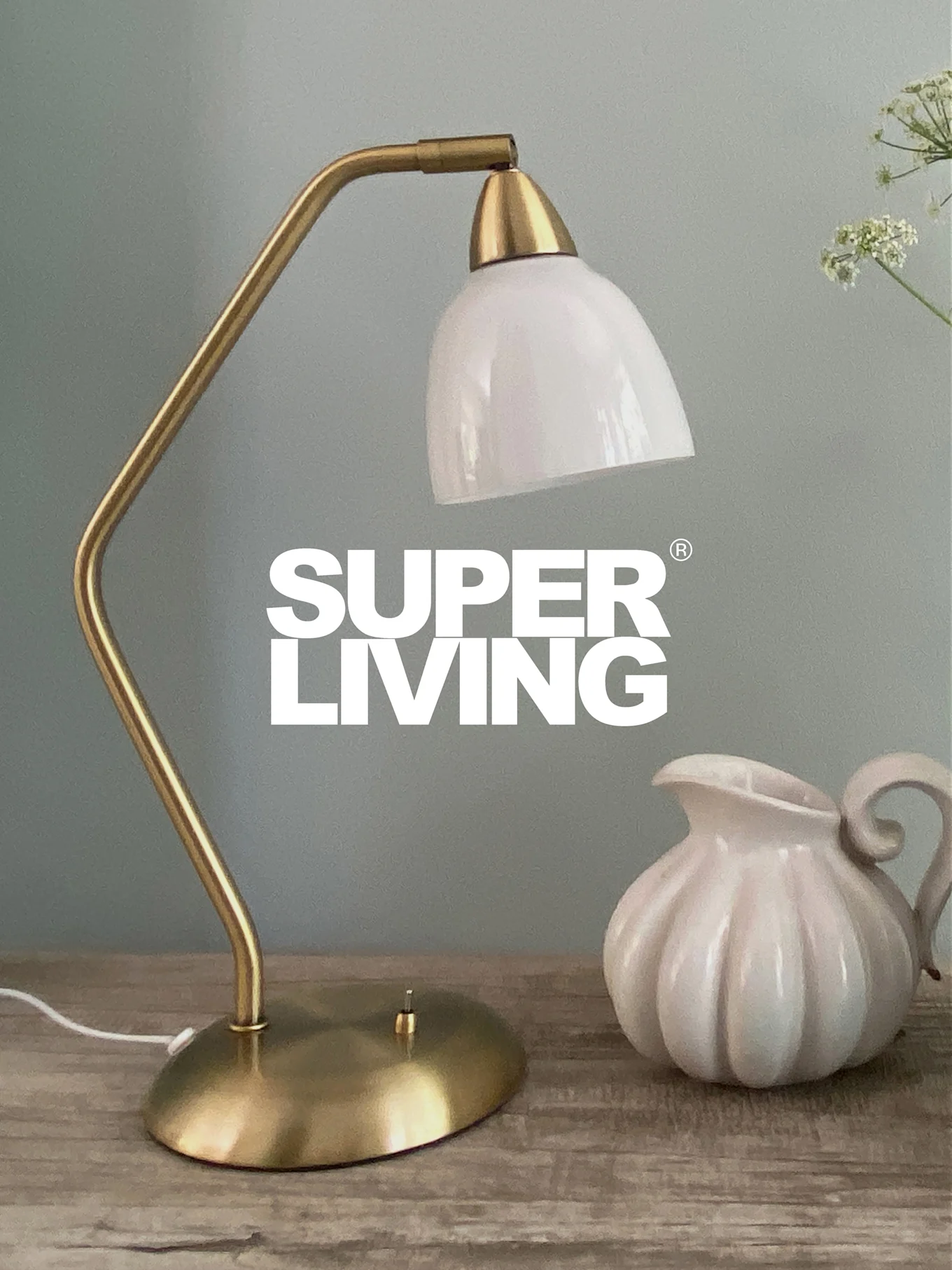 SUPER LIVING branding over en messingbordlampe med hvid skærm og en hvid, riflet kande på et træbord.