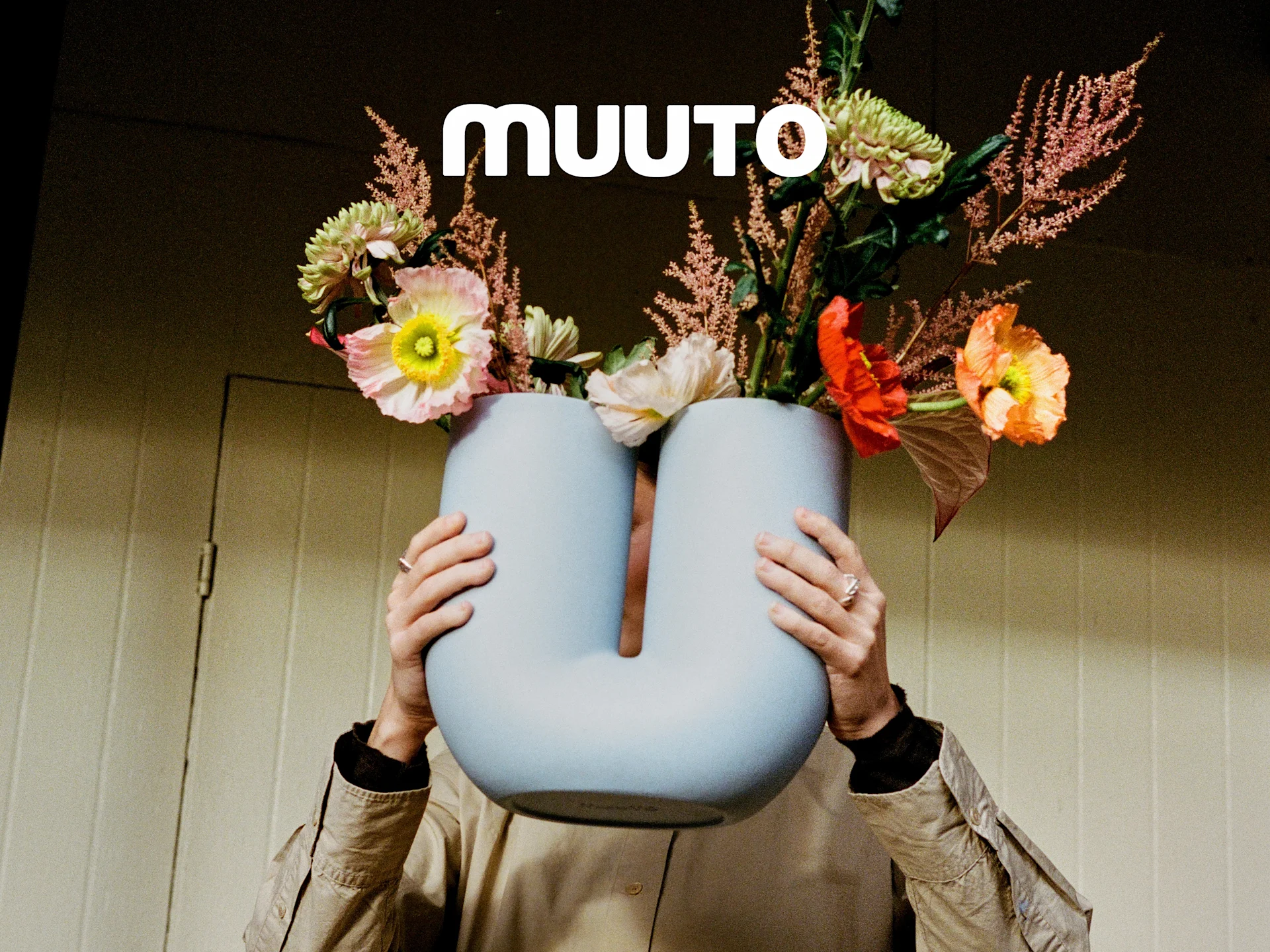 Person holder en lyseblå, U-formet vase med en levende buket af lyserøde, orange og grønne blomster. Muuto logo.