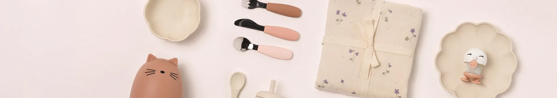 Flad lay af babyspisestel: beige tallerkener, katteflaske, lyserødt bestik, blomstret tæppe, æggeformet legetøj.